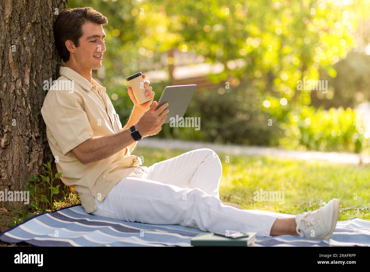 Relaxed guy sitting under tree at park, using digital tablet Stock Photo