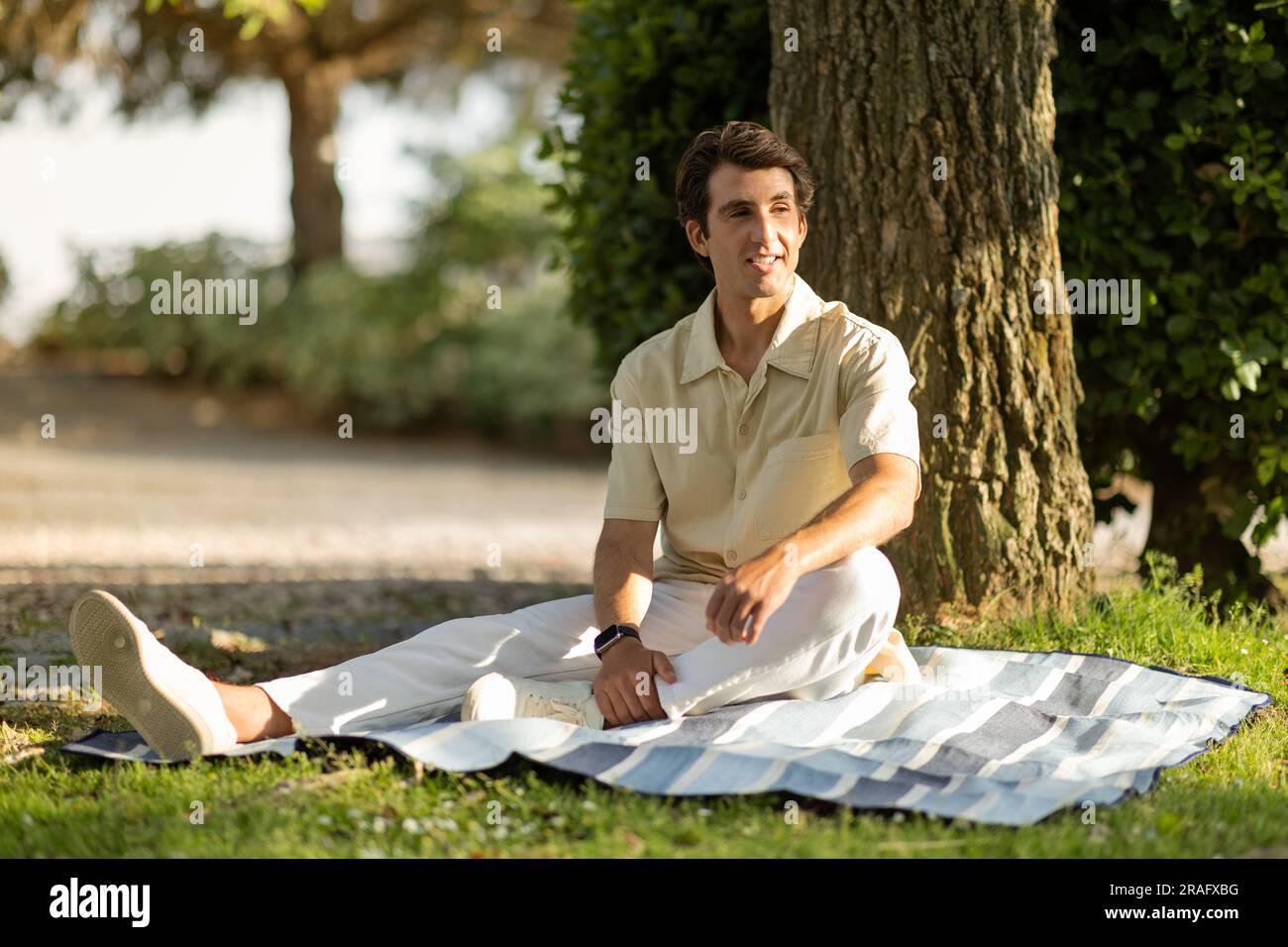 Positive handsome millennial man sitting under tree at park Stock Photo