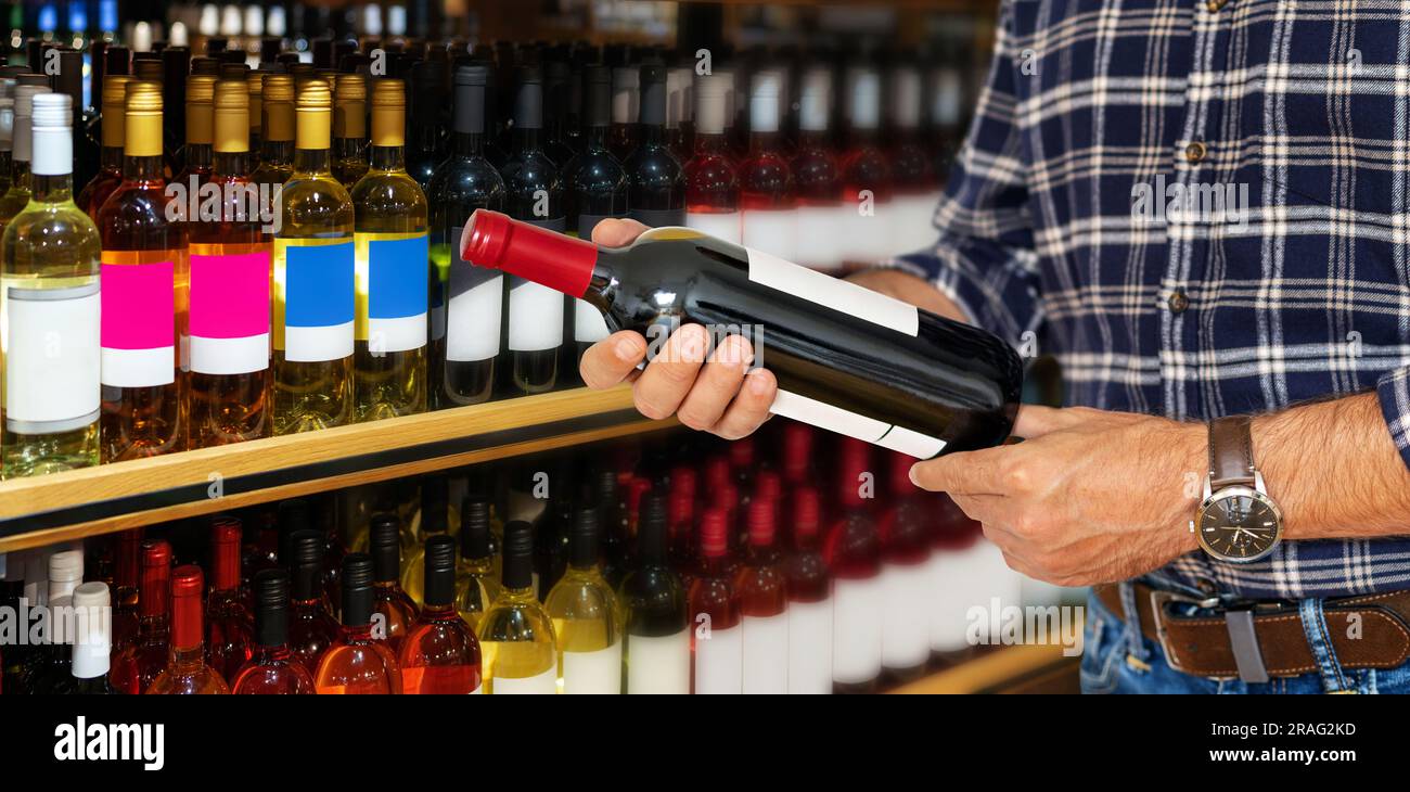 Customer in shop holding bottle of wine in his hands and reading information about the wine on the label. Stock Photo