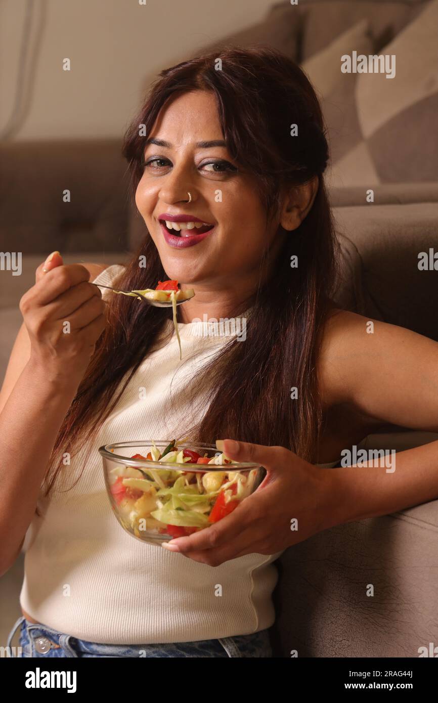 Happy Indian health conscious girl in living room having a bowl of green salad sitting besides a couch. Stock Photo