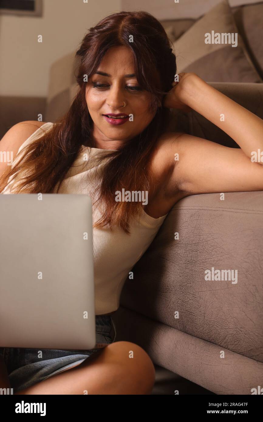 Happy Indian beautiful girl in living room working from home sitting besides a couch. Stock Photo