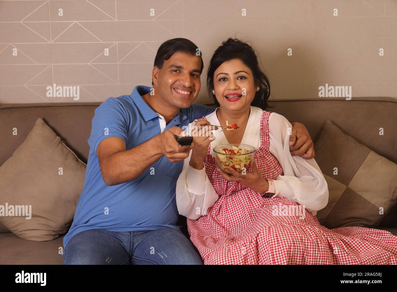 Happy Indian health conscious Husband wife sitting together having a bowl of green salad sitting on couch in a living room Stock Photo