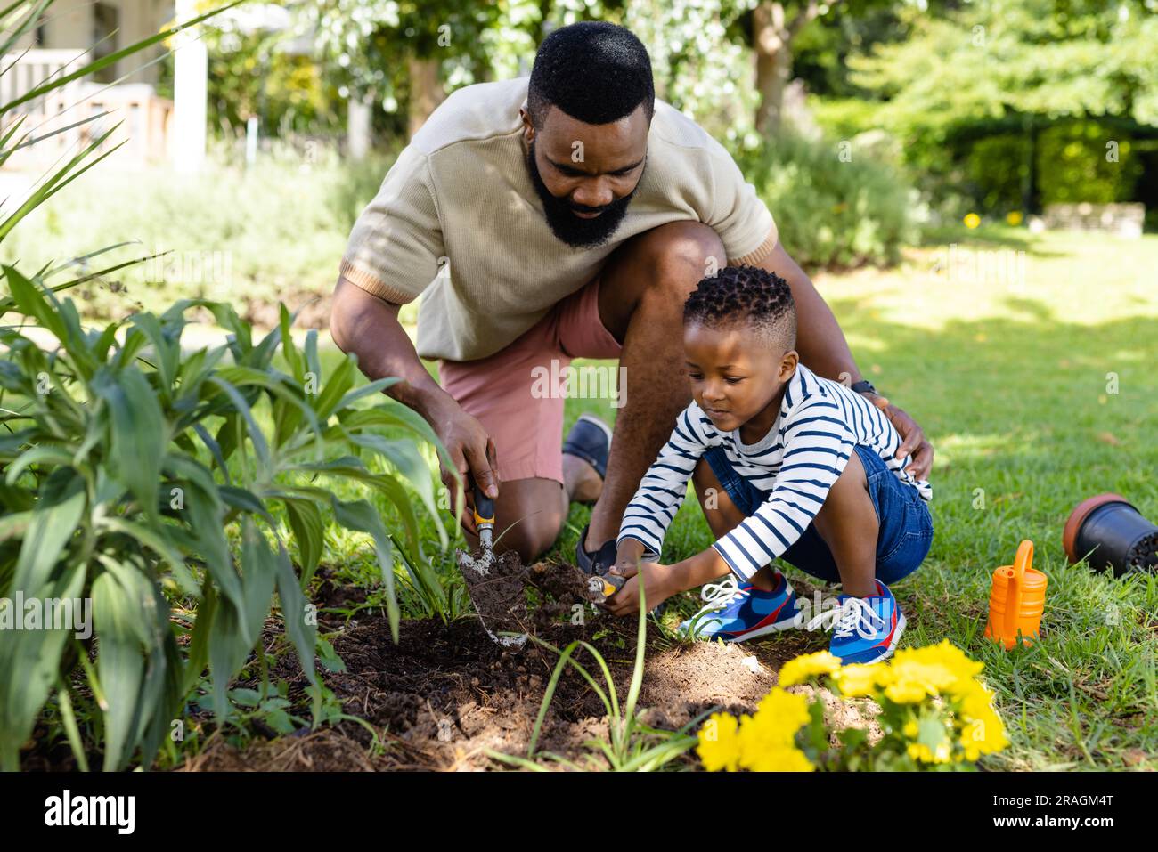 African american father assisting son in digging dirt with tools on grassy field in backyard Stock Photo