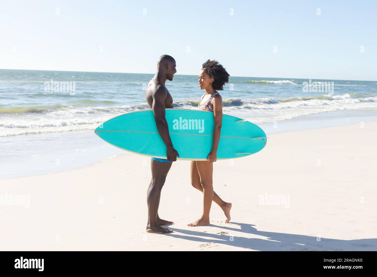 Happy african american couple holding surfboard smiling at each other standing on sunny beach Stock Photo