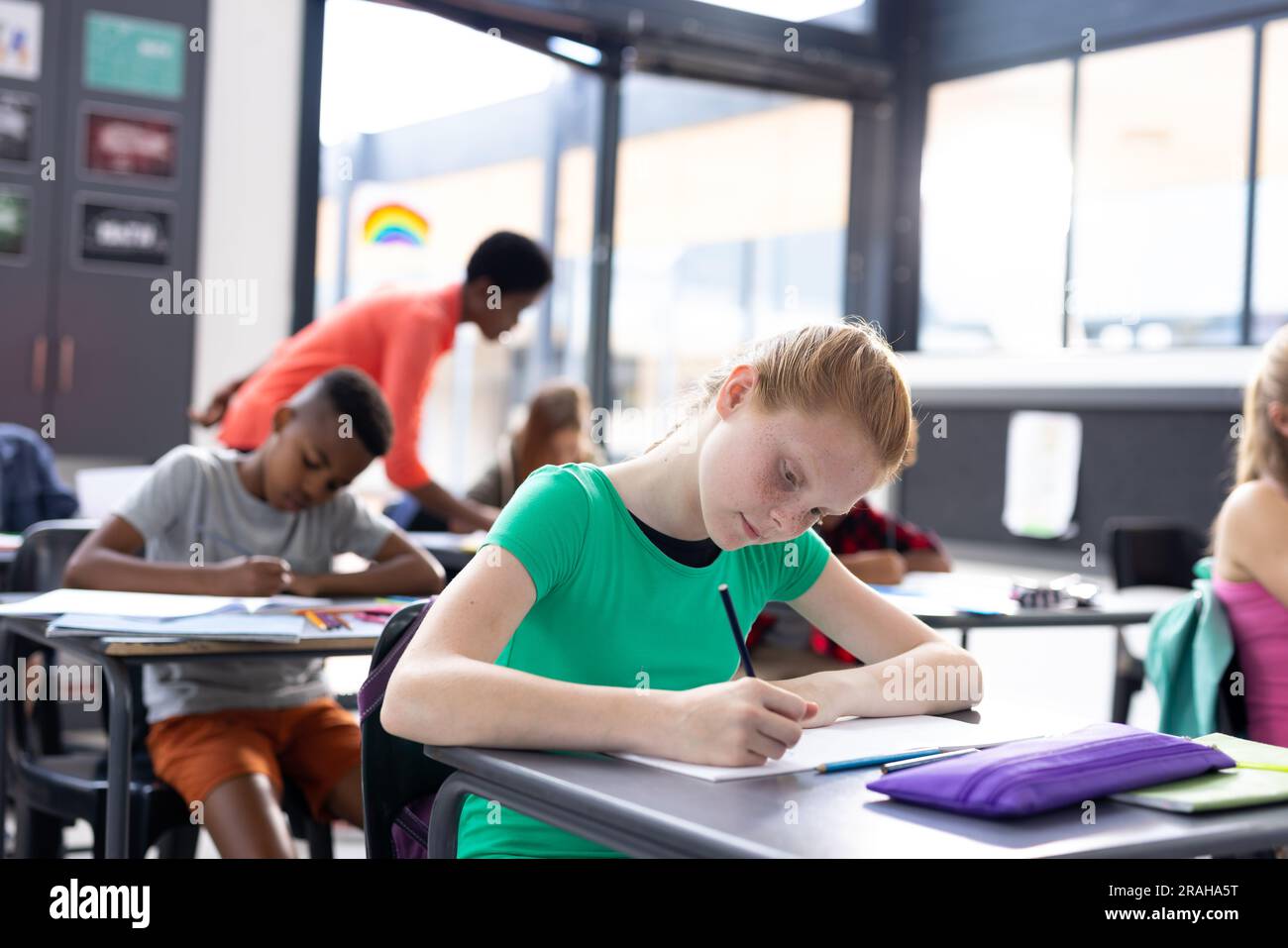 Happy caucasian schoolgirl writing at her desk with diverse pupils and ...