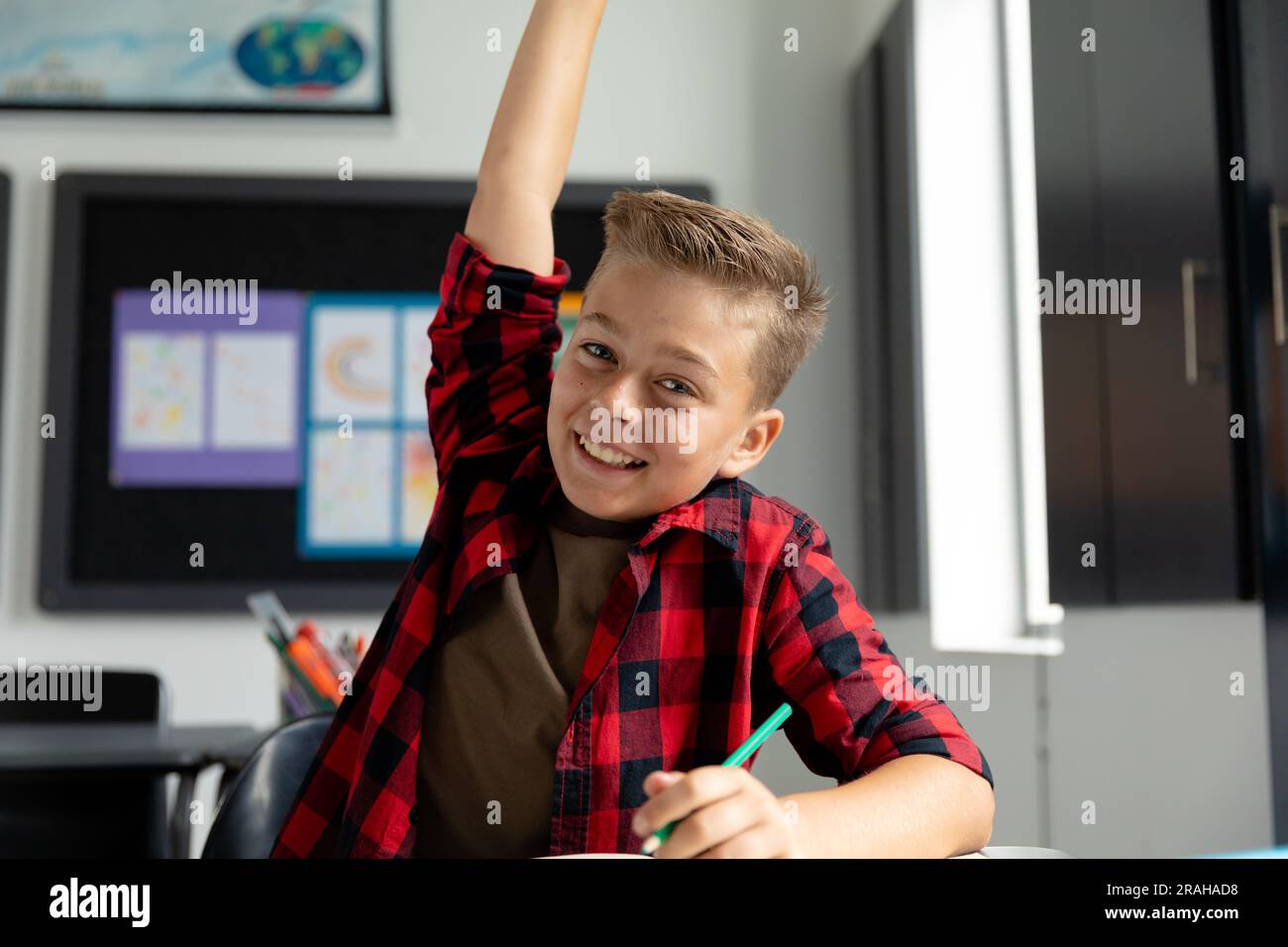 Eager, smiling caucasian schoolboy sitting at desk raising hand in ...