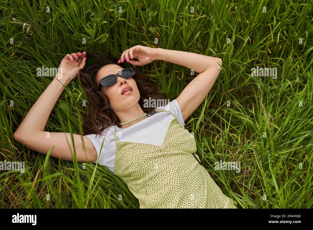 Top view of stylish brunette woman in sunglasses and sundress lying on grassy meadow and relaxing, peaceful retreat  and relaxing in nature concept, r Stock Photo