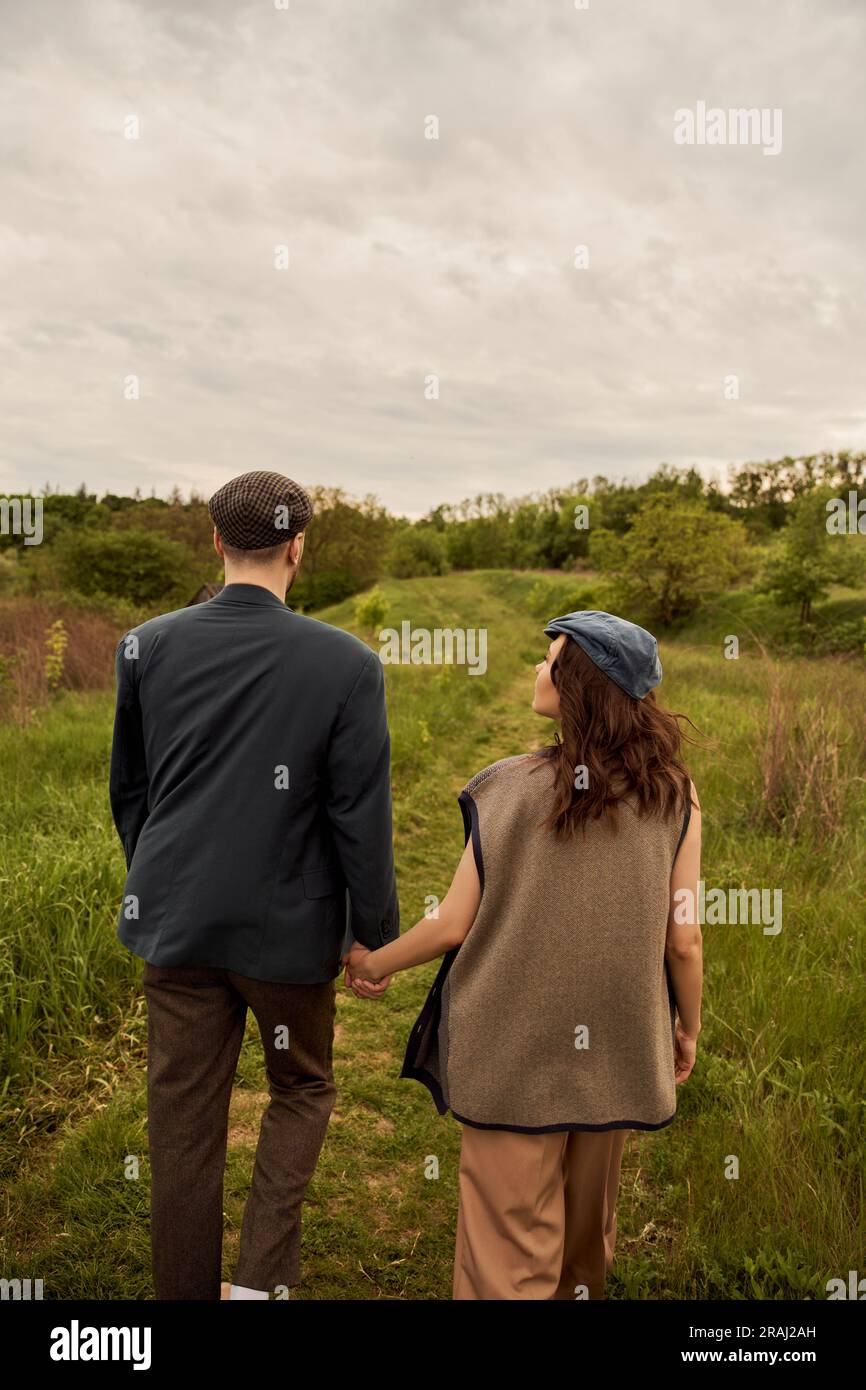 Brunette and fashionable woman in vest and newsboy cap holding hand of ...