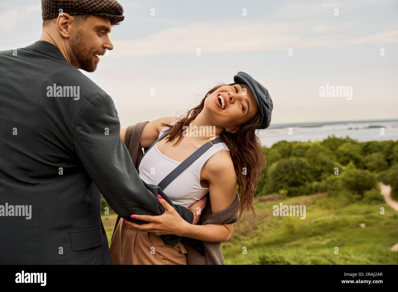 Smiling and bearded man in jacket and newsboy cap hugging cheerful ...