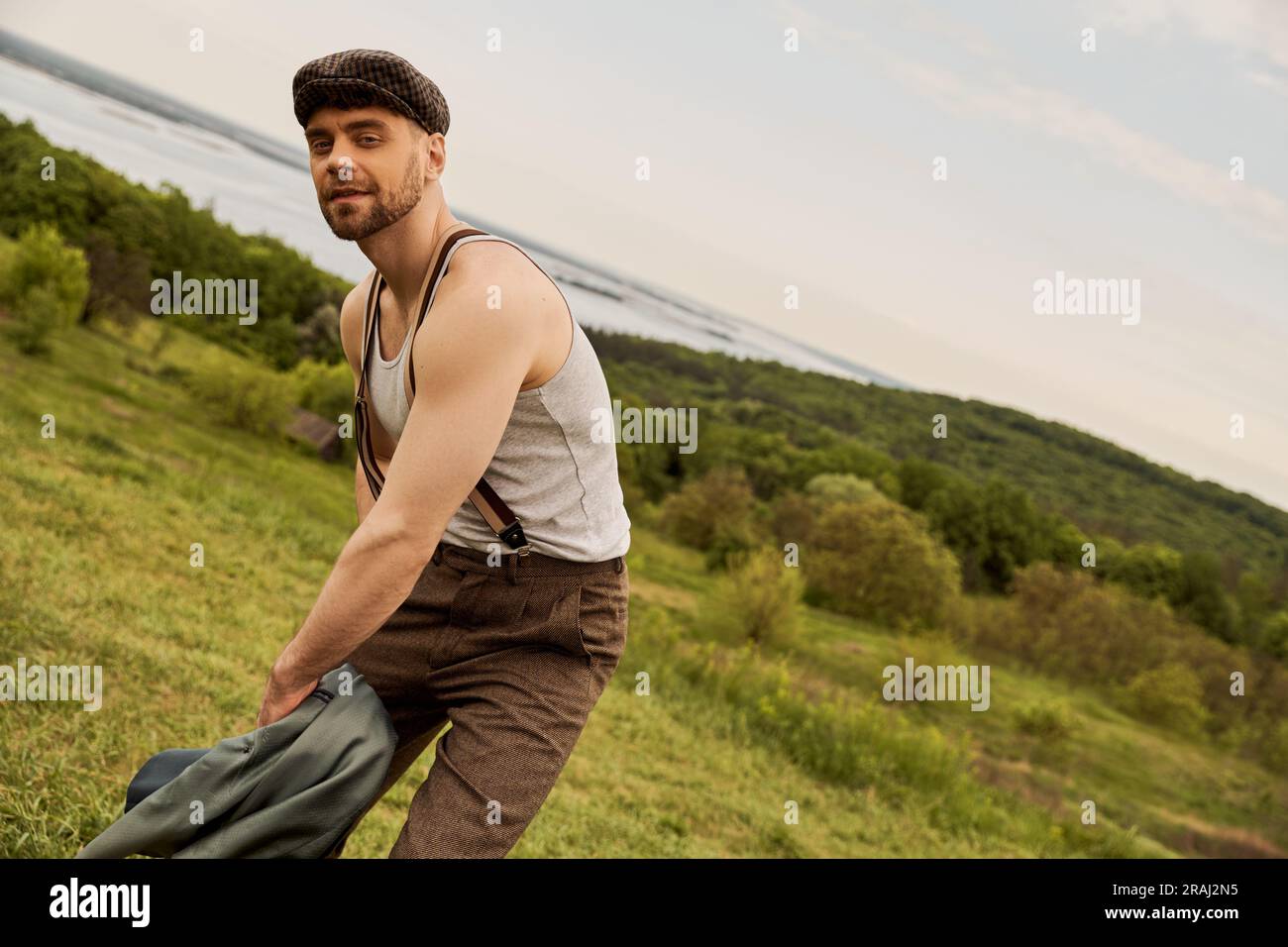 Trendy bearded man in newsboy cap and suspenders holding jacket and ...
