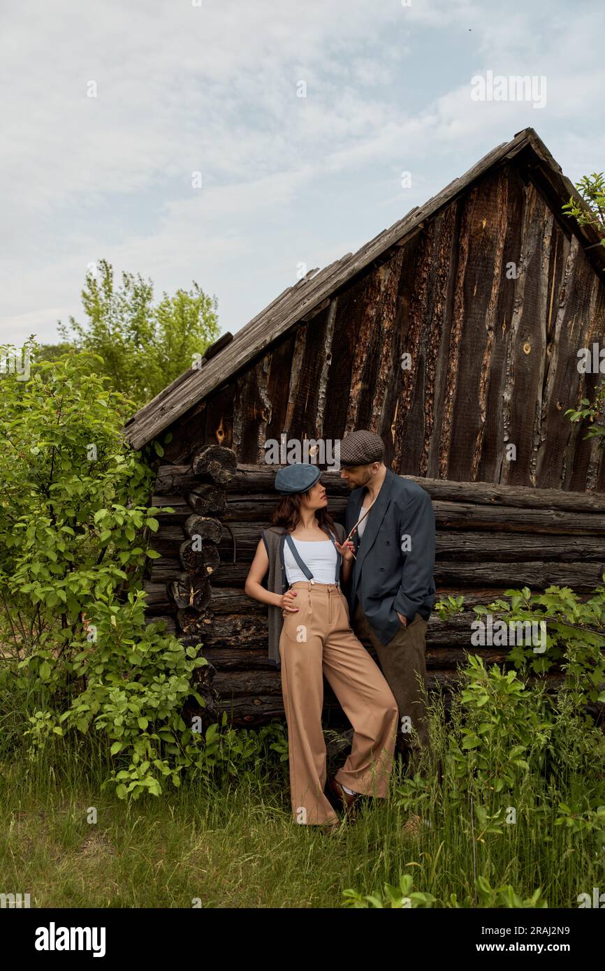 Fashionable woman in vintage outfit touching suspender while standing and looking at bearded boyfriend in newsboy cap near rural house on meadow, fash Stock Photo