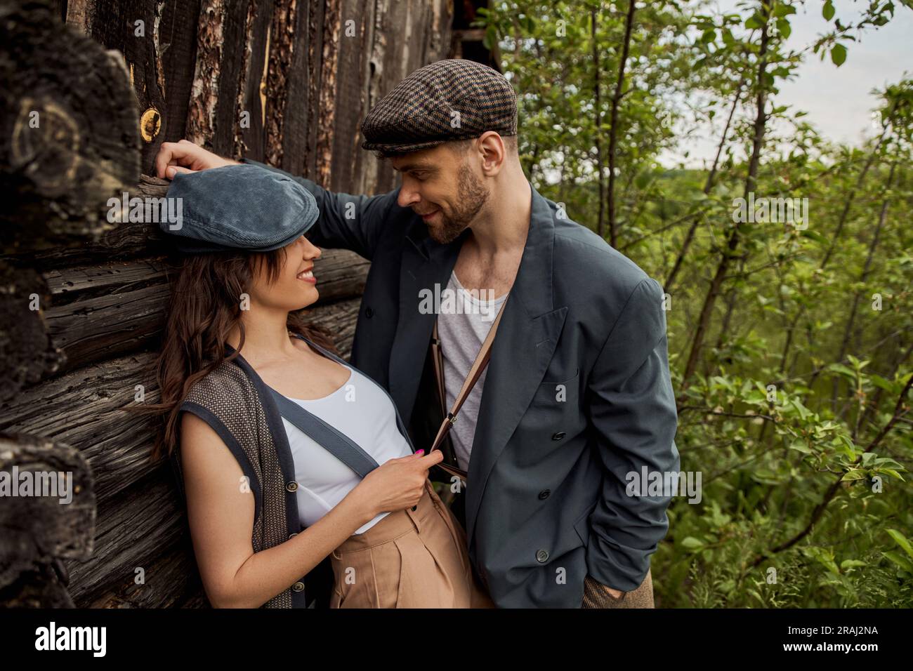 Cheerful and fashionable woman in newsboy cap touching suspender on bearded boyfriend in jacket while standing near rustic house with nature at backgr Stock Photo