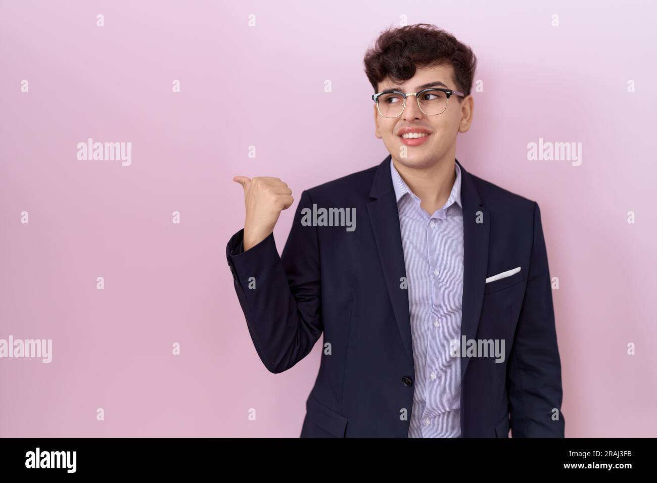 Young non binary man with beard wearing suit and tie smiling with happy face looking and pointing to the side with thumb up. Stock Photo