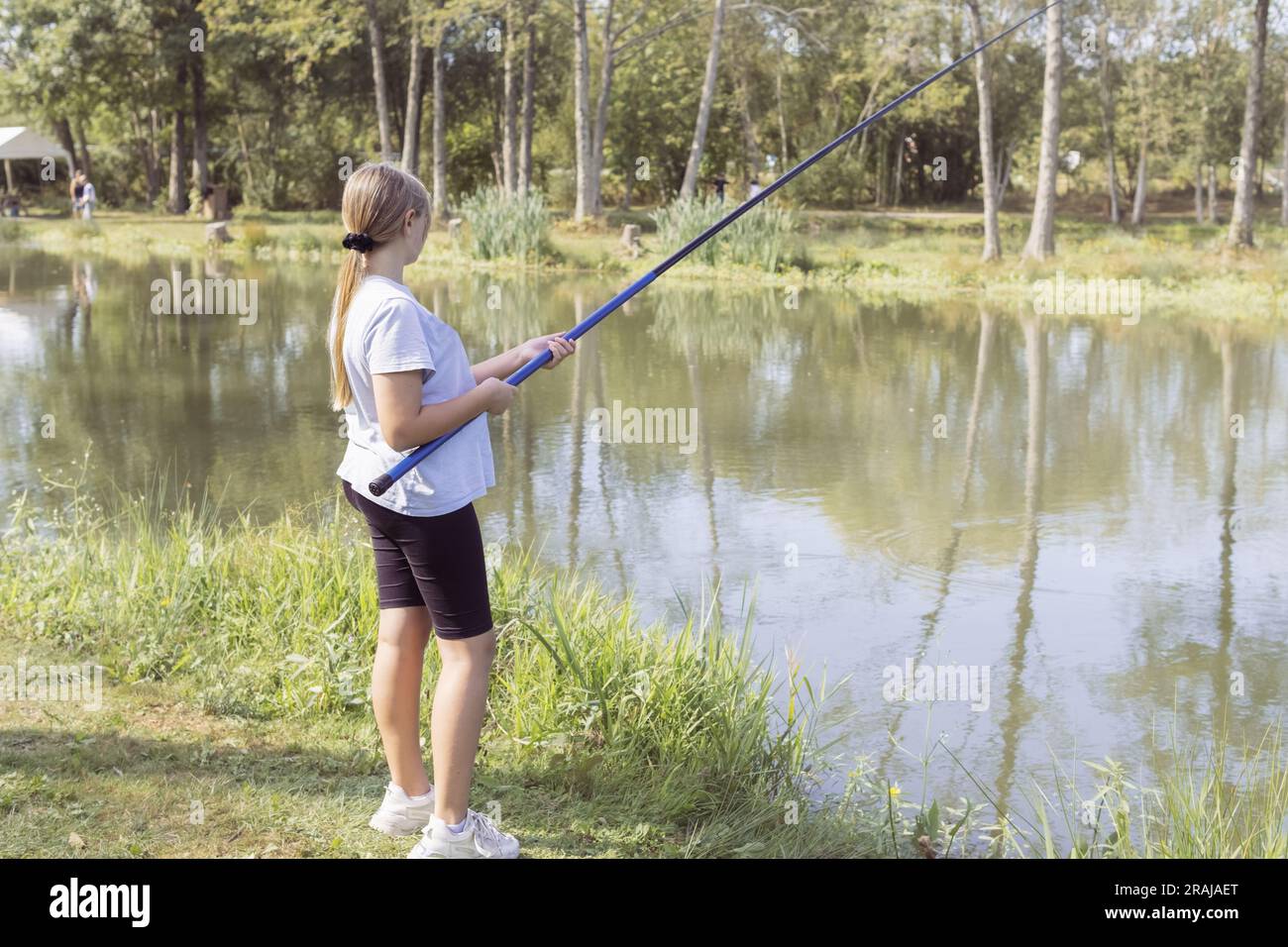 https://c8.alamy.com/comp/2RAJAET/a-teenager-girl-of-european-appearance-with-blond-hair-tied-in-a-ponytail-stands-fishing-at-a-stake-holds-a-fishing-rod-in-her-hands-dressed-in-bla-2RAJAET.jpg