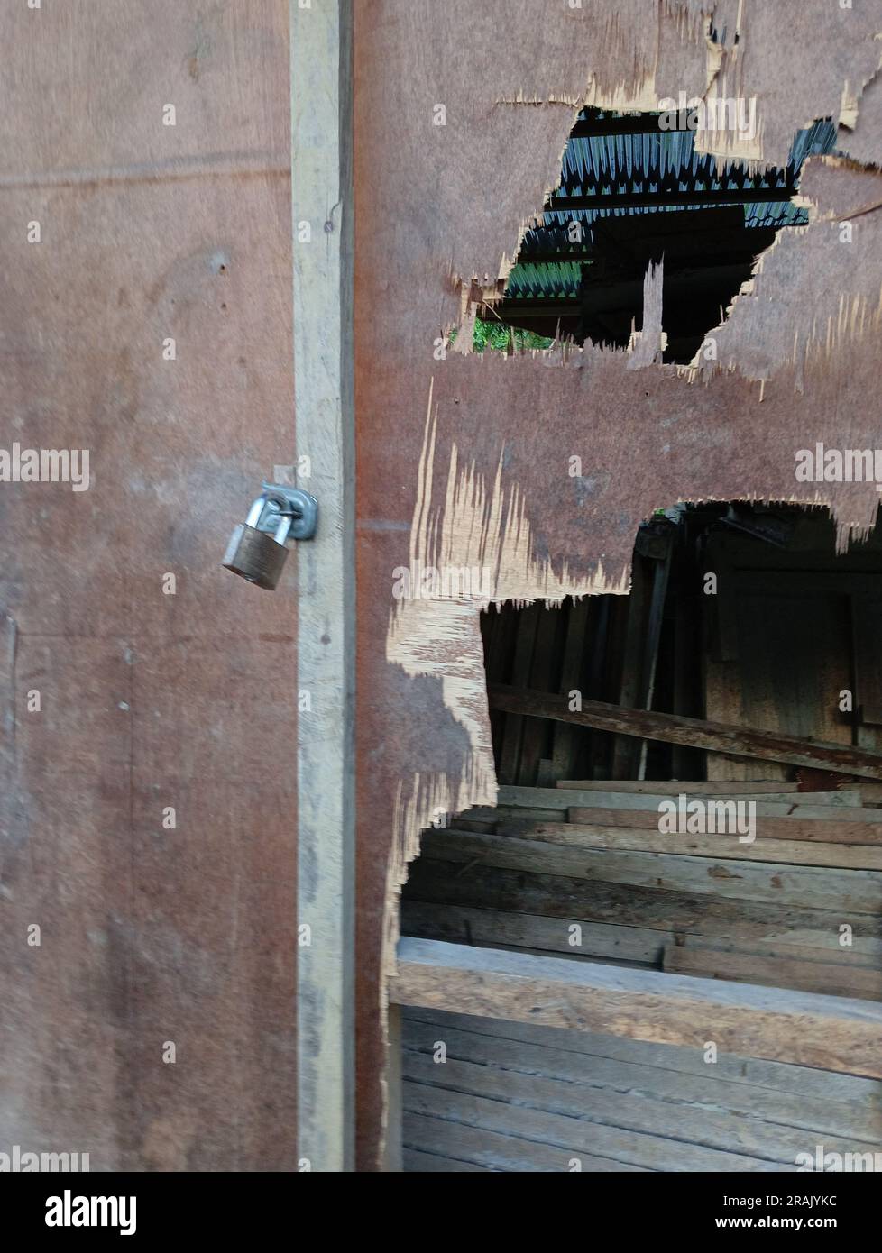 A small padlock attached to a door and wall made of plywood that has been damaged and has holes and shows the contents in the room. Stock Photo