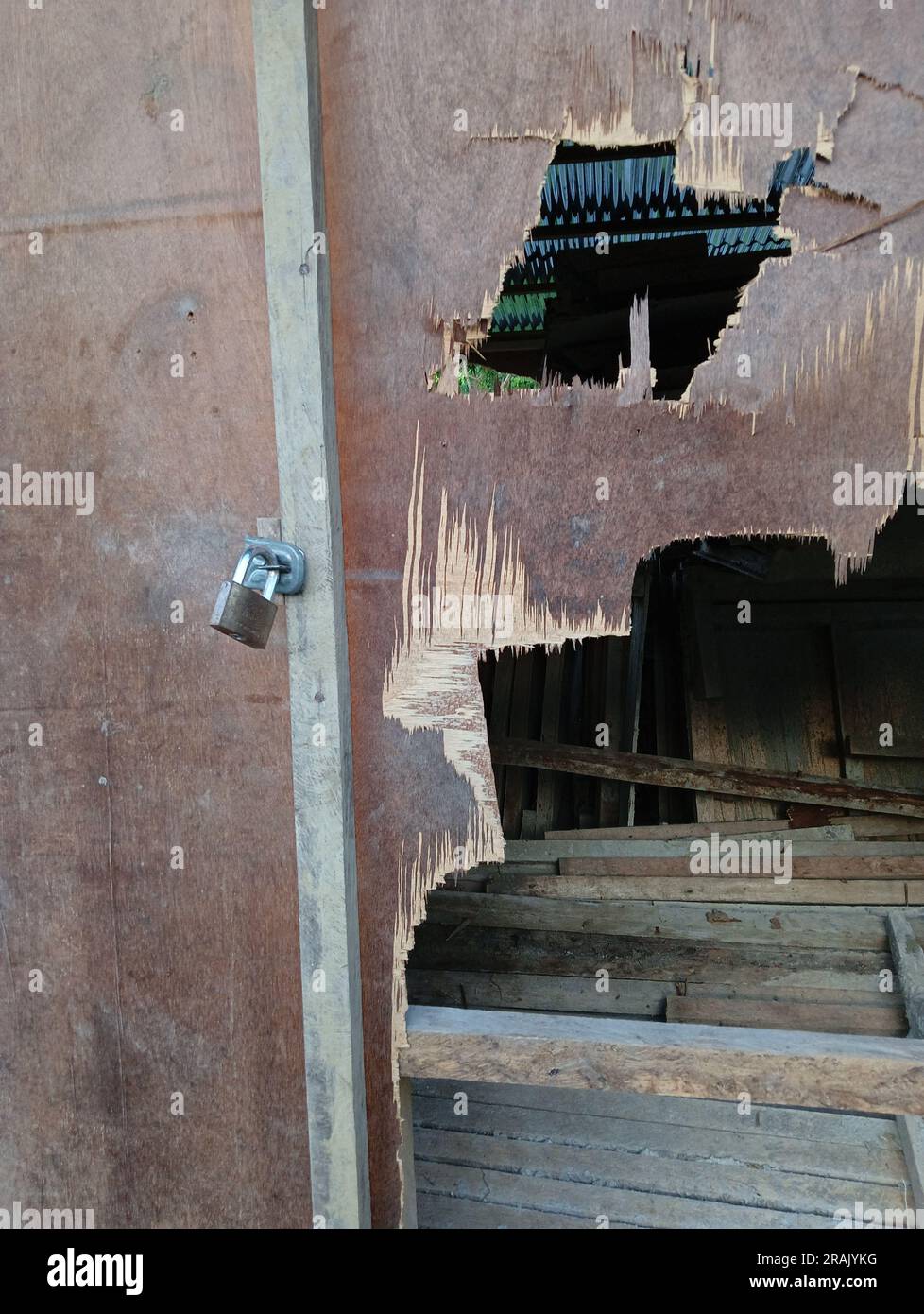 A small padlock attached to a door and wall made of plywood that has been damaged and has holes and shows the contents in the room. Stock Photo