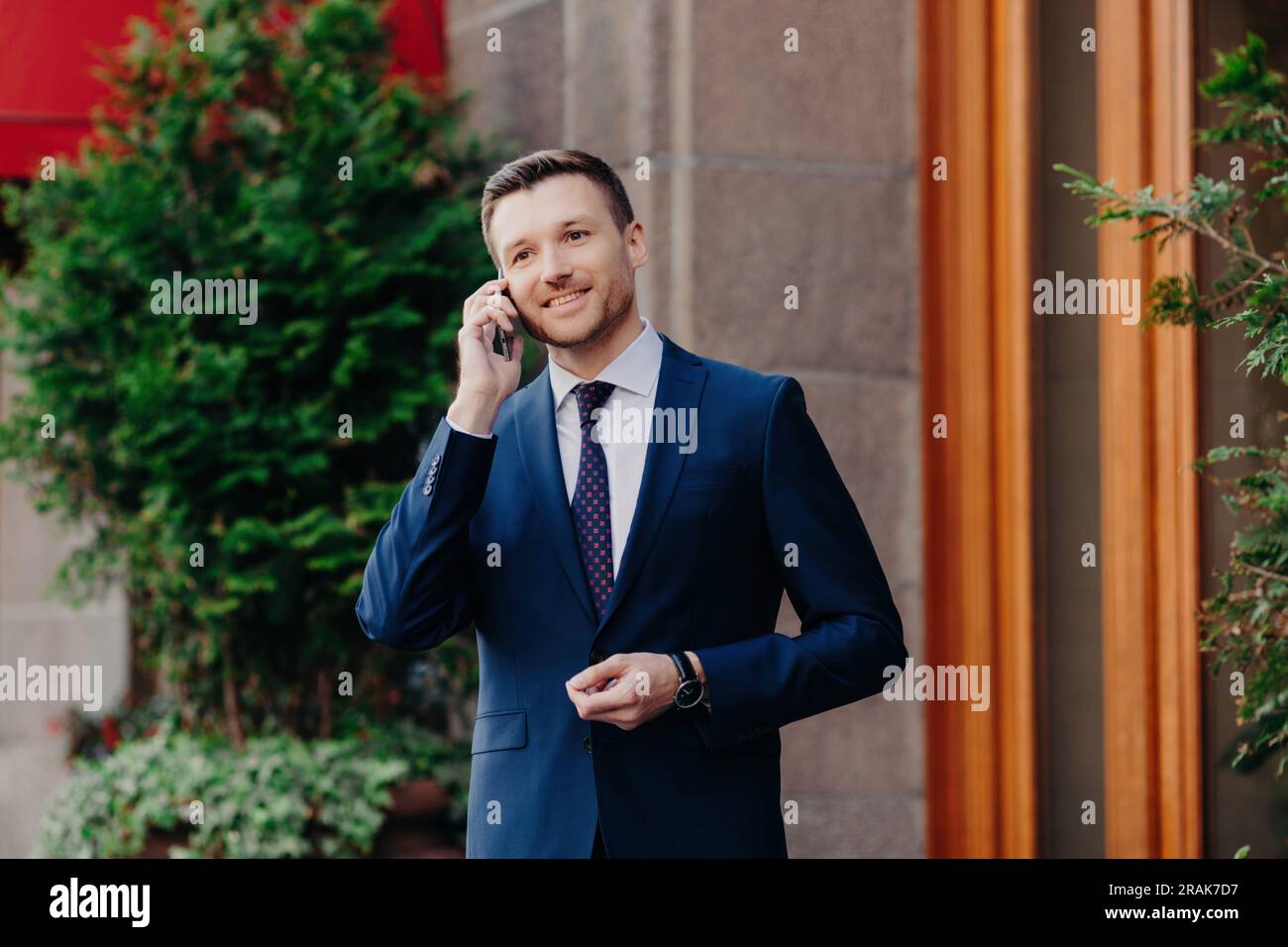 Busy businessman takes break, talks on phone, always connected. Formal attire, pleasant smile. Near restaurant. Tech-savvy. Stock Photo