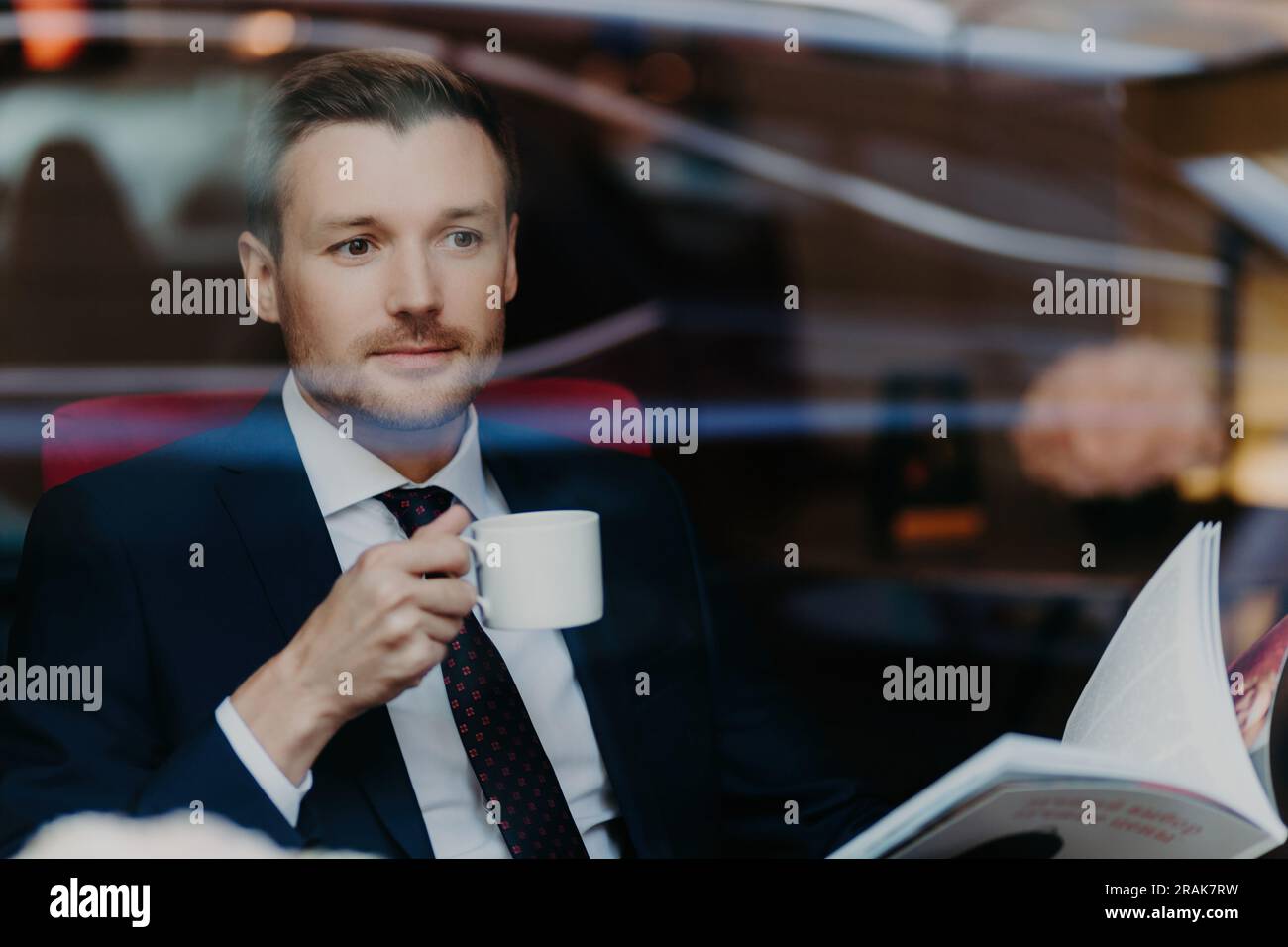 Handsome boss in luxury restaurant, holds cup, looks through menu, formal suit, deep in thought Stock Photo