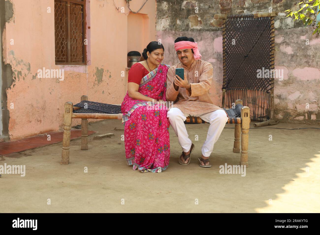 Portrait of Indian rural farmer couple sitting outdoors together in Indian villager's attire holding phone in hand. Stock Photo
