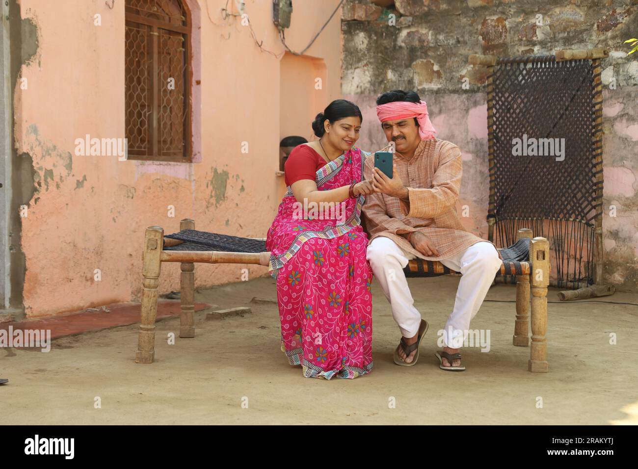 Portrait of Indian rural farmer couple sitting outdoors together in Indian villager's attire holding phone in hand. Stock Photo