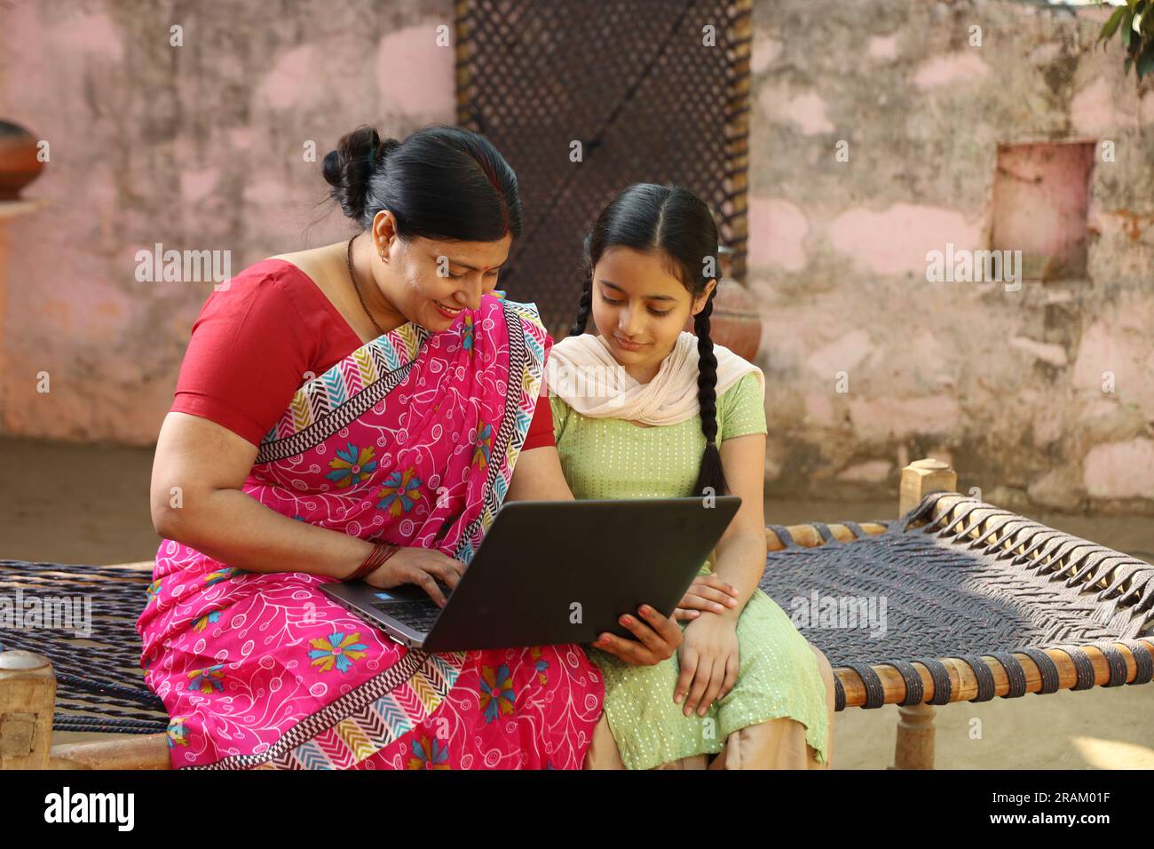 Happy Indian village family of single mother and daughter using laptop sitting outside the house. Digital India. Child education. Woman empowerment. Stock Photo