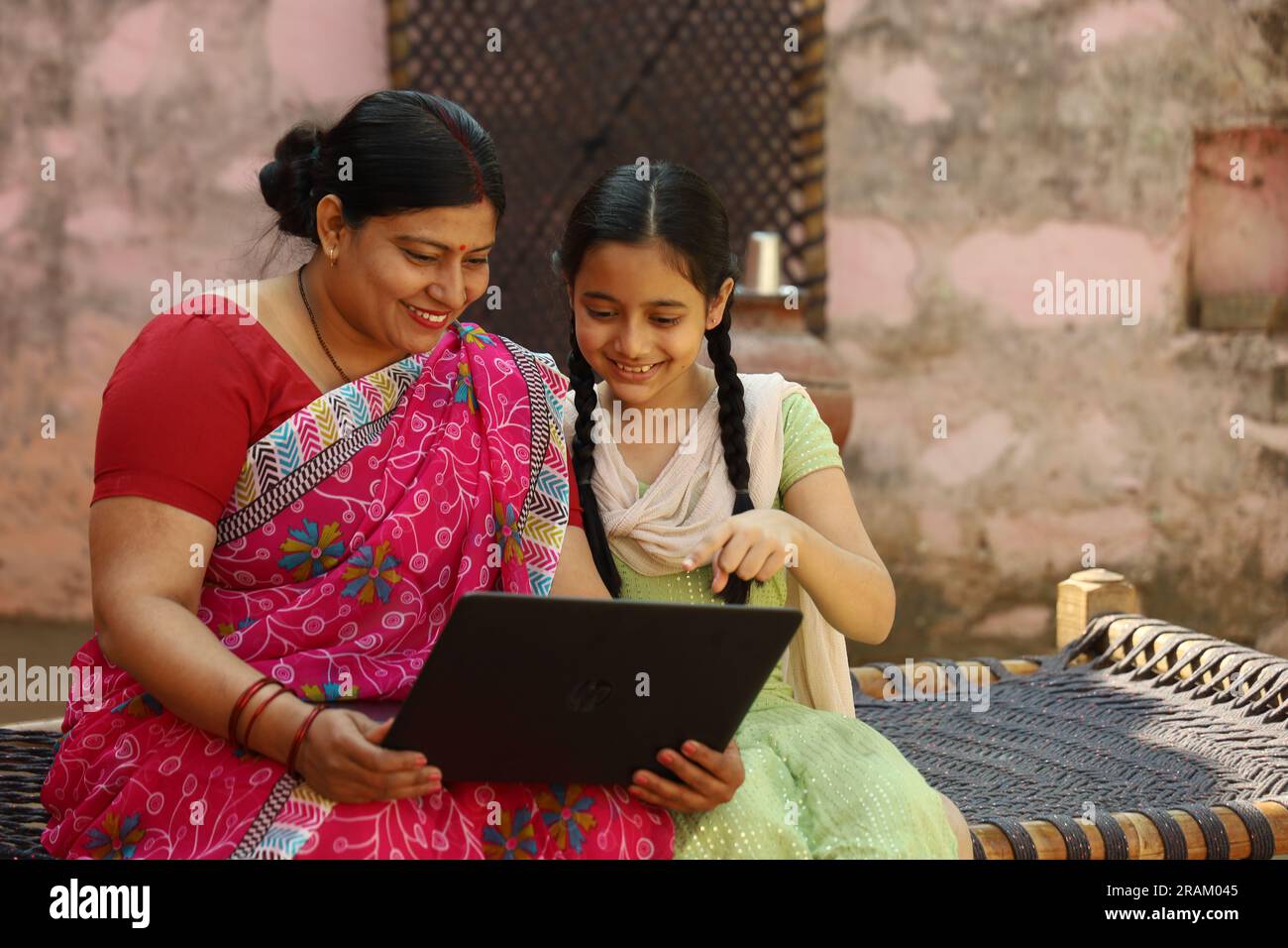 Happy Indian village family of single mother and daughter using laptop sitting outside the house. Digital India. Child education. Woman empowerment. Stock Photo