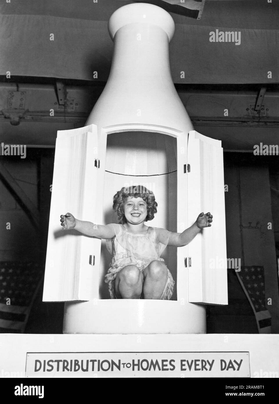 Chicago, Illinois:  c. 1930 A young girl with curly hair and open arms appears in an oversize milk bottle displayed at a milk promotional booth in a tradeshow. Stock Photo