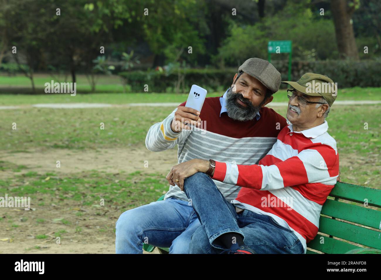 Old father and mid aged son sitting on park bench holding mobile phone in hand. Video call and taking selfie. They are in lush greens and serenity. Stock Photo