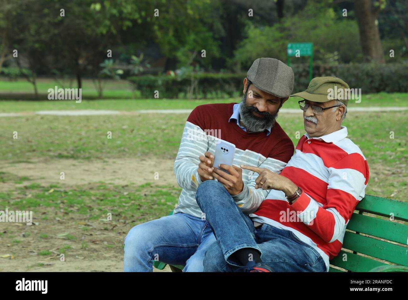 Old father and mid aged son sitting on park bench holding mobile phone in hand. Video call and taking selfie. They are in lush greens and serenity. Stock Photo