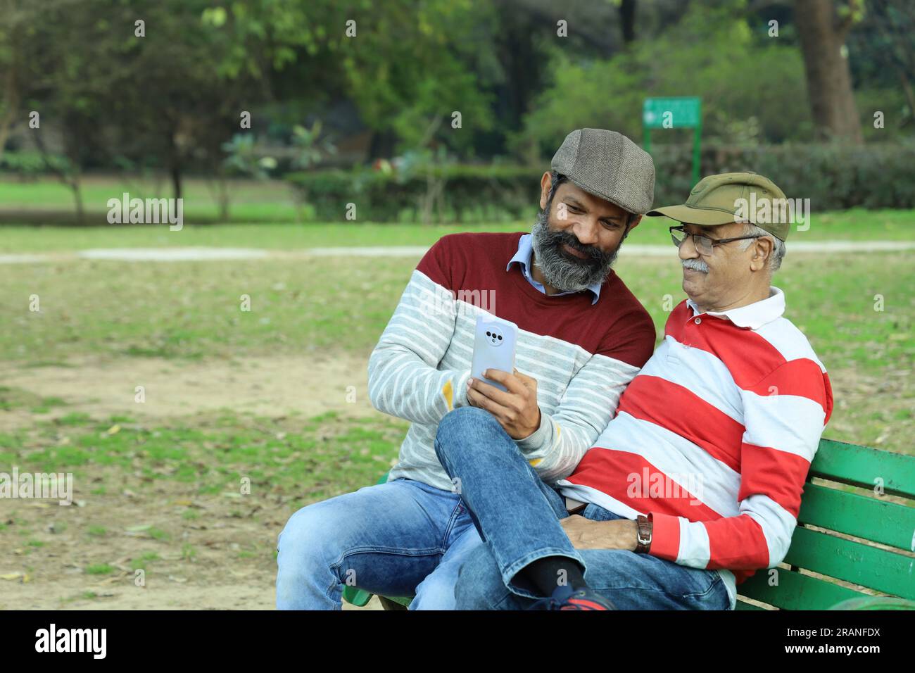 Old father and mid aged son sitting on park bench holding mobile phone in hand. Video call and taking selfie. They are in lush greens and serenity. Stock Photo