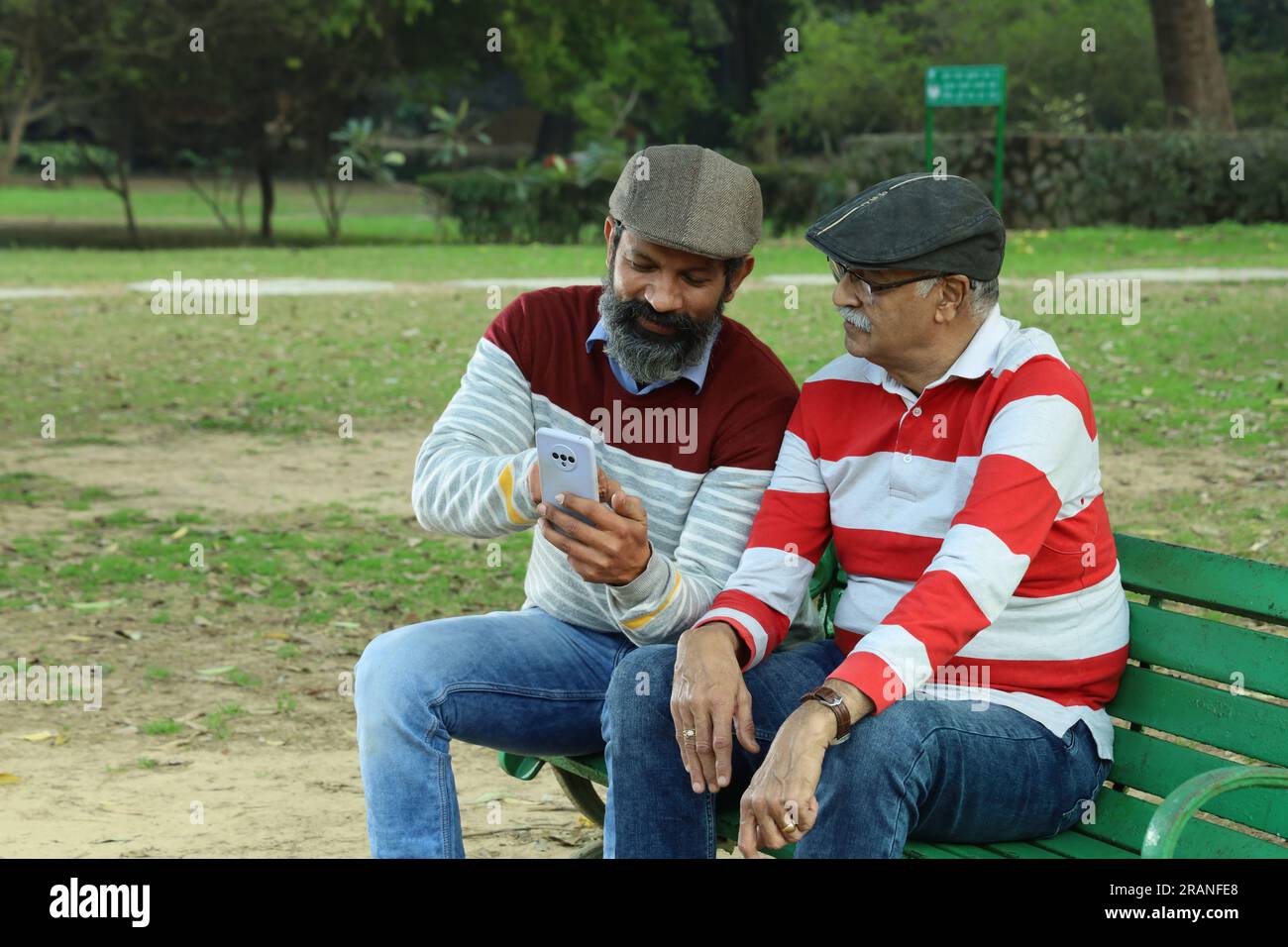 Old father and mid aged son sitting on park bench holding mobile phone in hand. Video call and taking selfie. They are in lush greens and serenity. Stock Photo