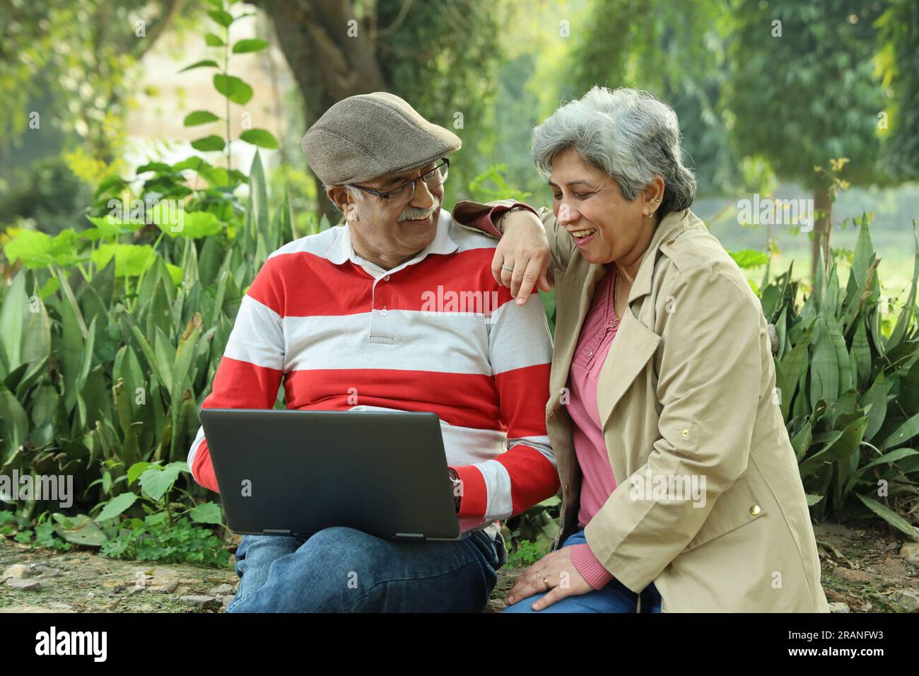 Old couple sitting in serene atmosphere in park holding laptop and working online, making best use of technology and internet to book their vacations. Stock Photo
