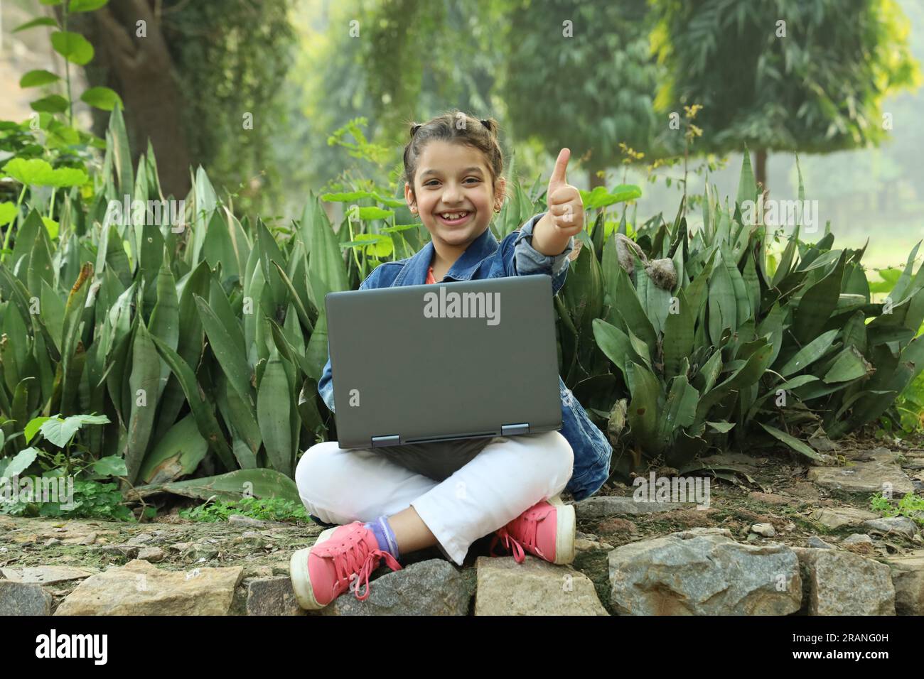 Young girl sitting in lush green atmosphere in park holding laptop and studying online, making best use of technology and internet. Study from home. Stock Photo
