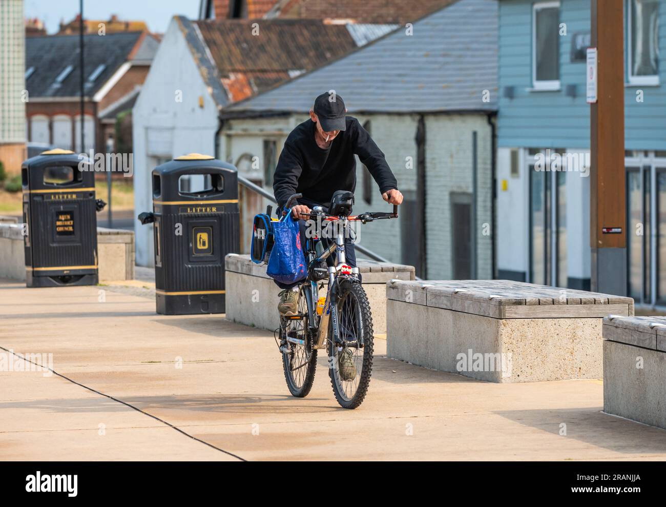 Male cyclist wearing a hat riding bicycle along promenade while smoking with cigarette hanging out his mouth, in the UK. Stock Photo