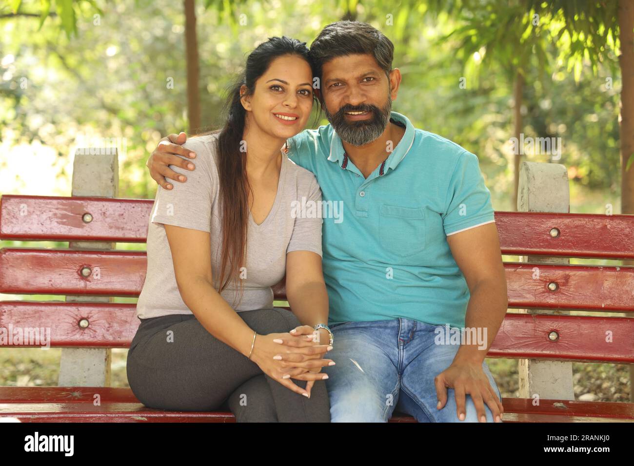 Portrait of a happy Indian couple sitting on the bench in the park amongst the greens. Stock Photo