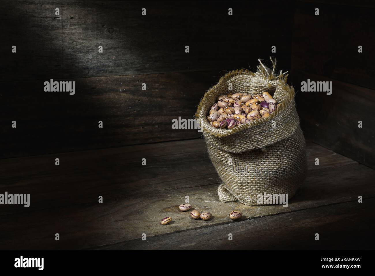 A still life of a pile of raw, dry, uncooked, beige, brown Borlotti beans in a hessian bag in the corner of a wooden box, in pools of soft mood light Stock Photo
