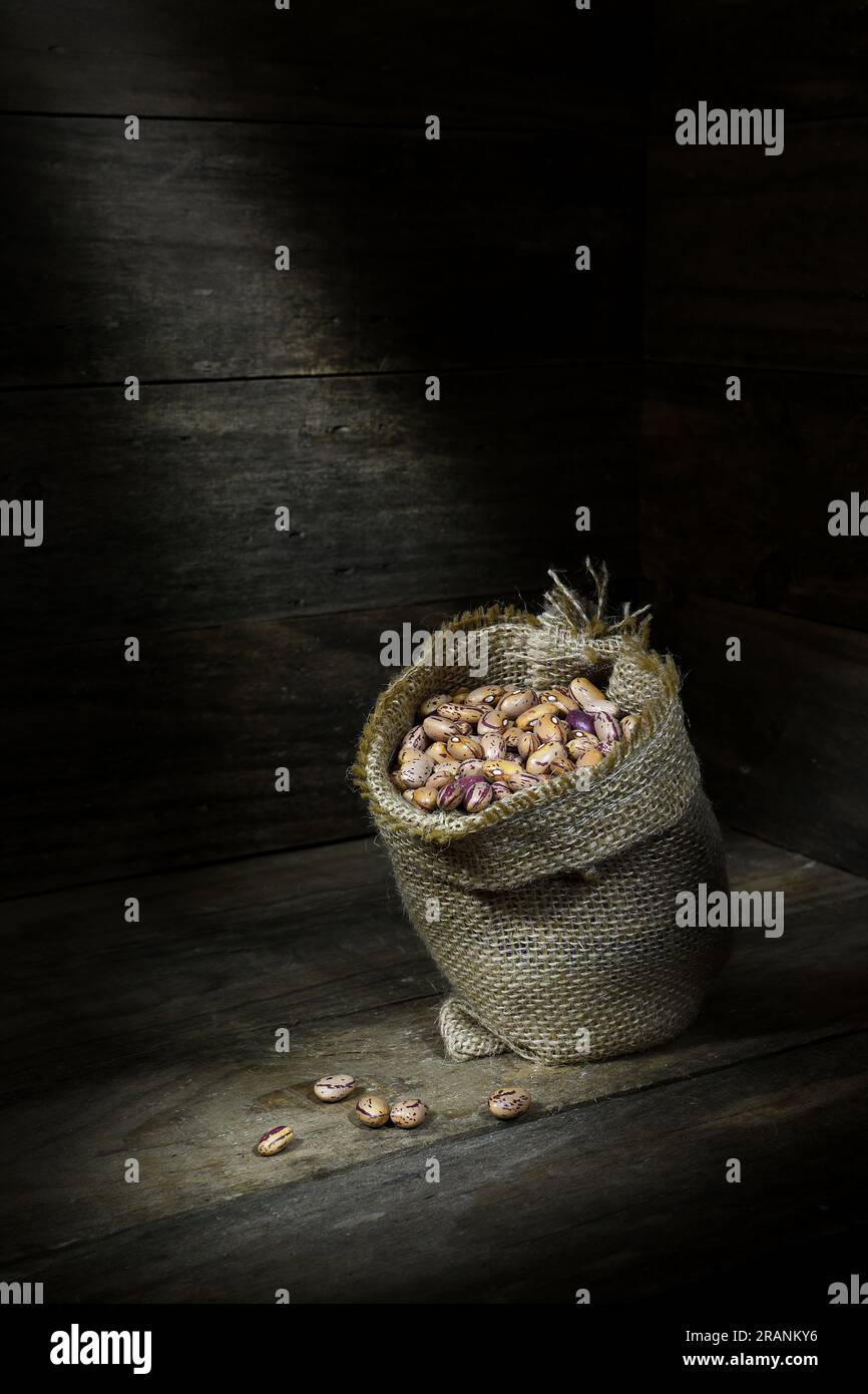 A still life of a pile of raw, dry, uncooked, beige, brown Borlotti beans in a hessian bag in the corner of a wooden box, in pools of soft mood light Stock Photo