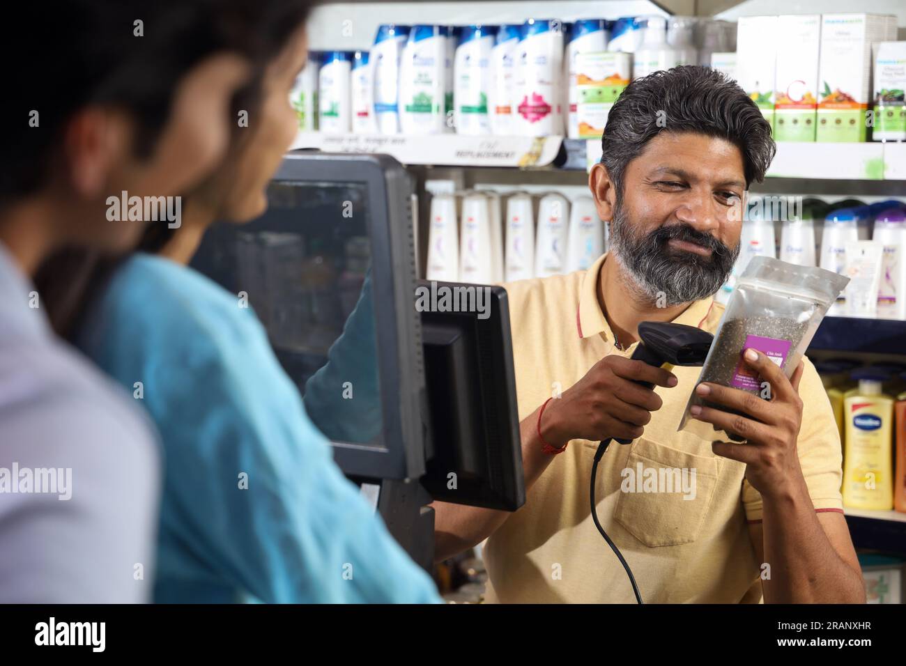 Bearded guy on the billing counter scanning the products. Customers standing in the queue in a mini shopping hyperstore. supermart. retail store. Bill Stock Photo