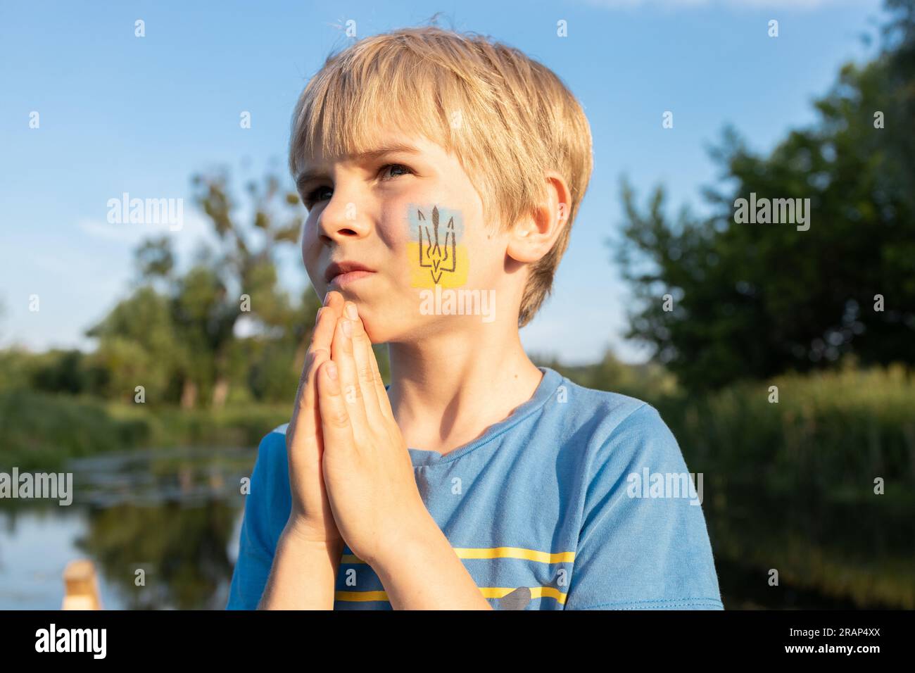 boy with painted trident and a Ukrainian flag on his cheek folded his hands in a prayer-appeal to God. Children against war. Ukrainian child asks for Stock Photo