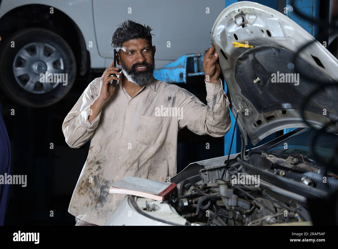 Close up portrait of bearded happy car mechanic standing in service station beside a car. Car Specialist talking on phone while examining the engine. Stock Photo