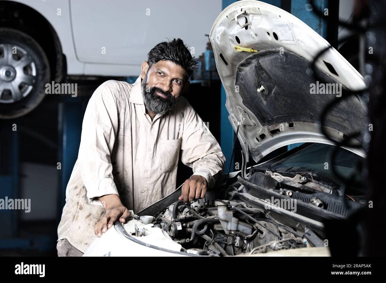 Close up portrait of bearded happy car mechanic standing in service station beside a car. Car Specialist is examining the engine. Stock Photo