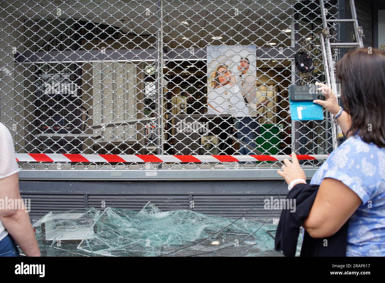A woman takes a photo with her phone. Optique, 7 Rue Louise Thuliez, Place de Fêtes, 75019, Paris, France, 30th June, 2023. Optique opticians is attacked after a third night of violence and riots over the police killing of a teenager on Tuesday evening in Nanterre a suburb of Paris. Credit: Jane Burke/Alamy Live News. Stock Photo