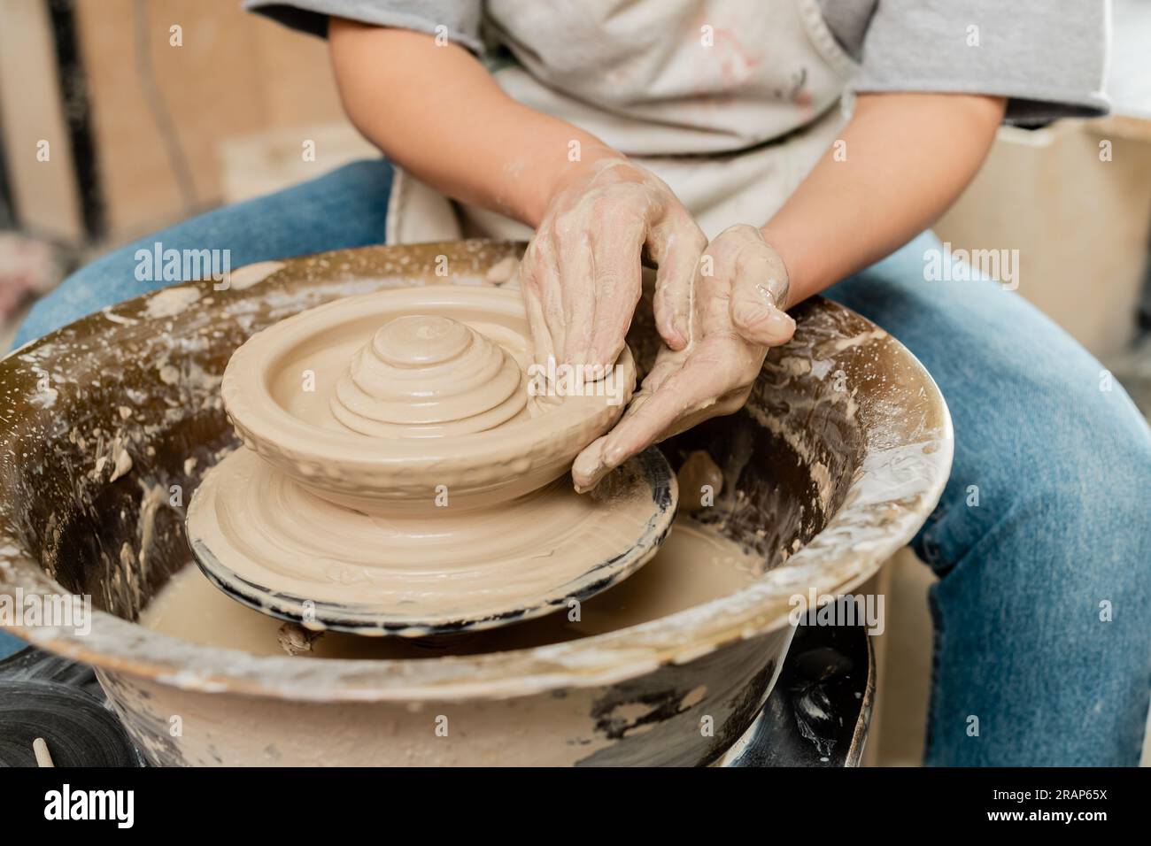 Cropped view of blurred female artisan in apron making shape of clay on spinning pottery wheel while working in pottery class, skilled pottery making Stock Photo