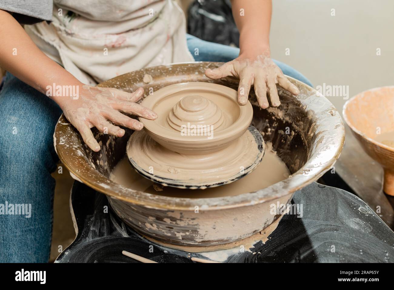Cropped view of young craftswoman in apron making shape of clay on spinning pottery wheel in blurred ceramic workshop at background, skilled pottery m Stock Photo