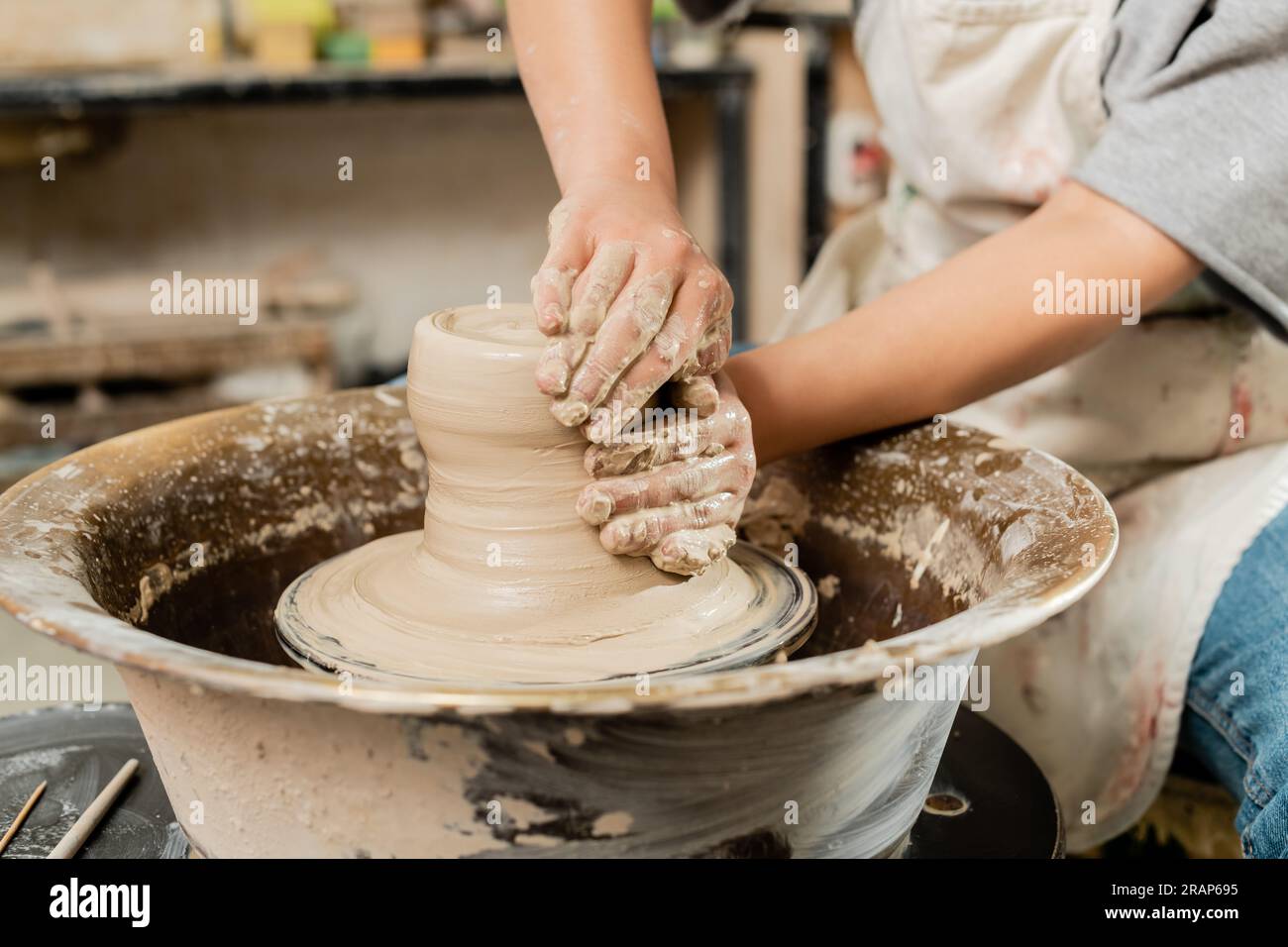 Cropped view of blurred female ceramicist in apron molding wet clay and working on spinning pottery wheel in art ceramic studio, skilled pottery makin Stock Photo