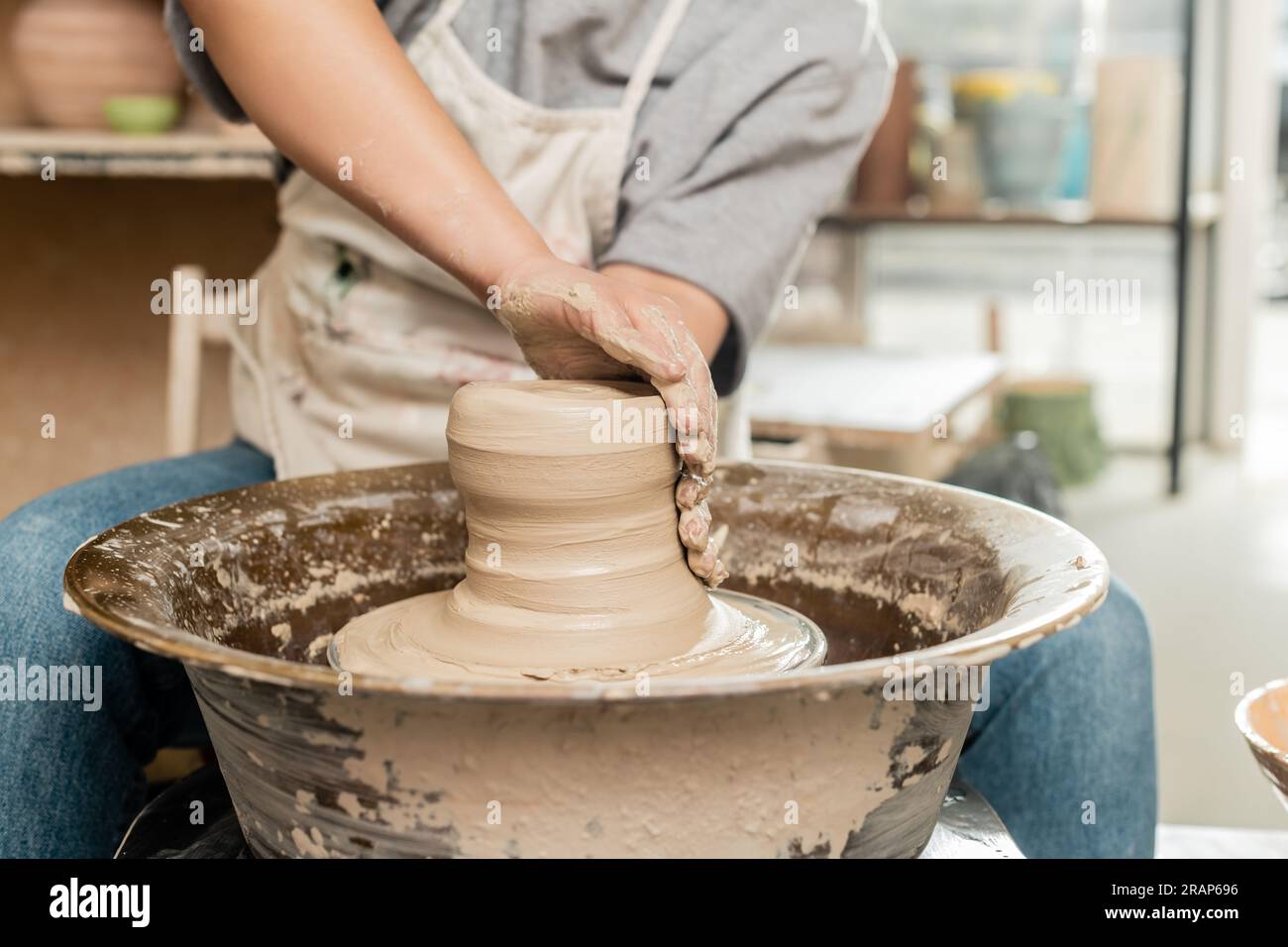 Cropped view of blurred female ceramicist in apron molding and shaping wet clay while working with spinning pottery wheel in art workshop, skilled pot Stock Photo