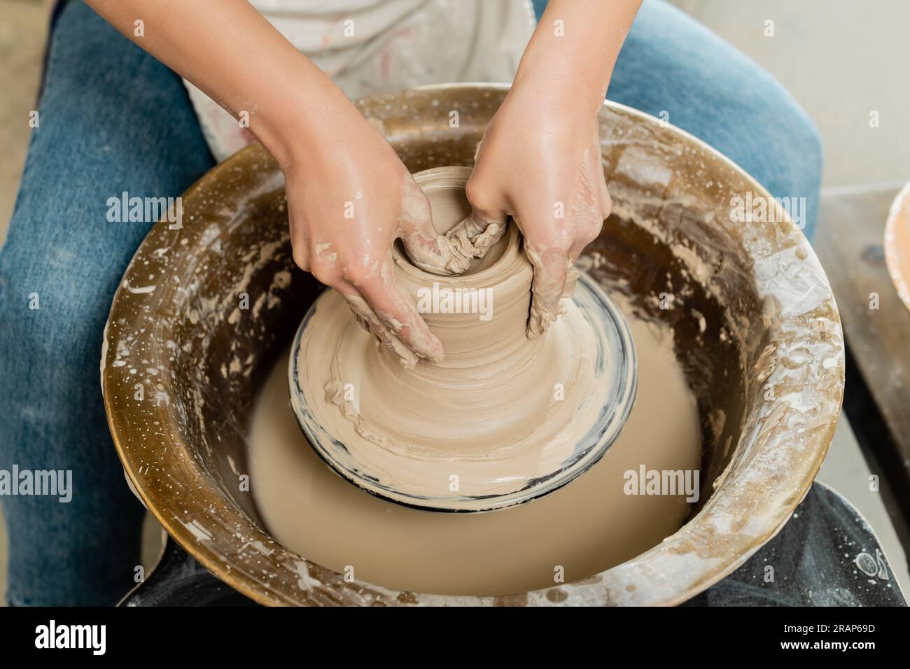High angle view of young female potter in apron shaping wet clay while working on spinning pottery wheel in art workshop at background, skilled potter Stock Photo