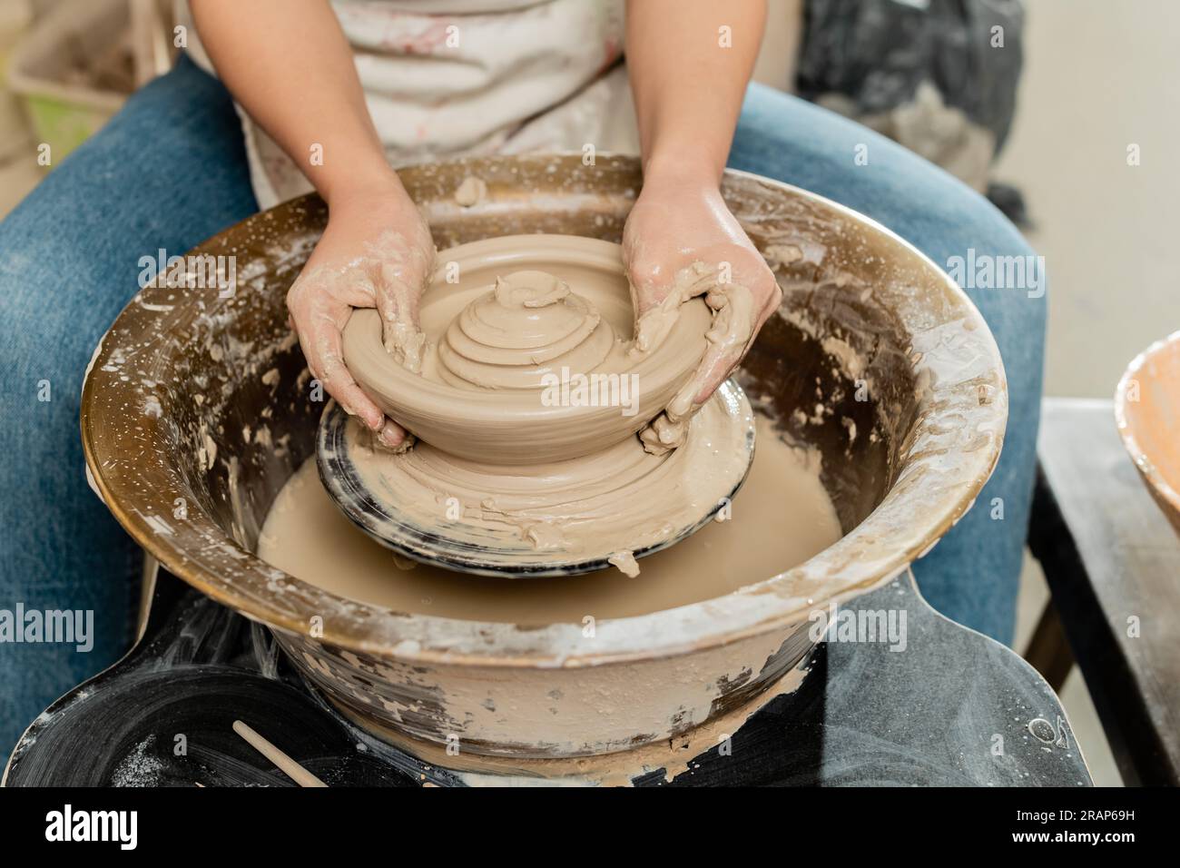 Cropped view of blurred female artisan in apron making shape of wet clay on spinning pottery wheel while working in ceramic workshop, skilled pottery Stock Photo