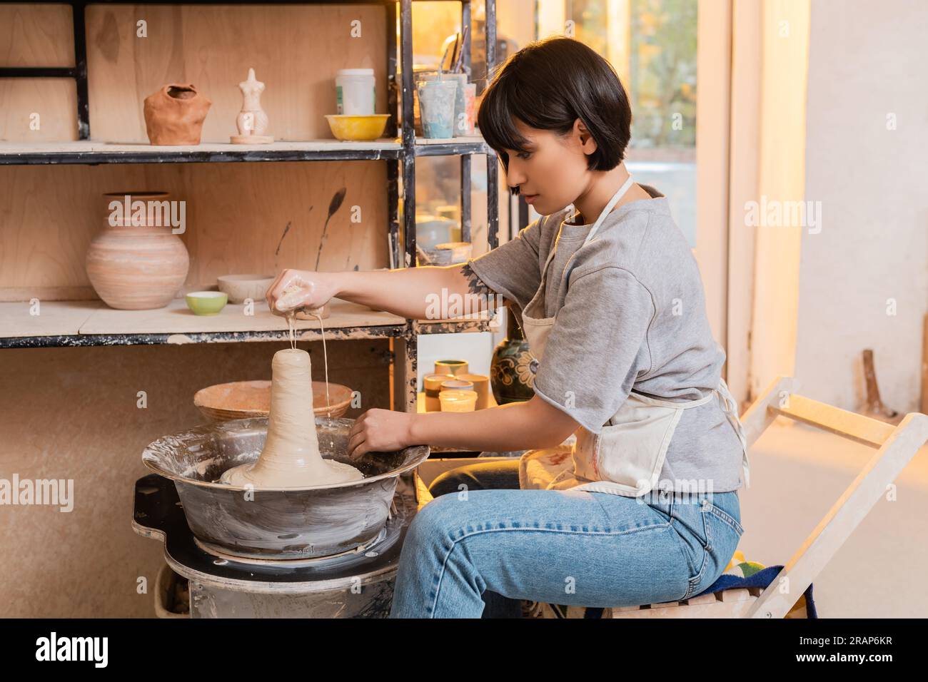 Side view of young asian female artisan in apron pouring water from sponge on wet clay on pottery wheel and working in ceramic workshop at sunset, pot Stock Photo