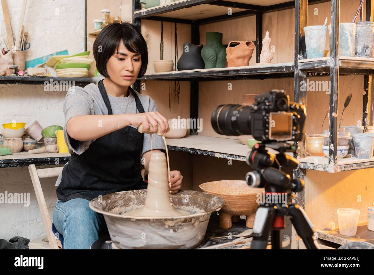 Young brunette asian female artisan in apron pouring water from sponge on wet clay and pottery wheel near digital camera on tripod in studio at backgr Stock Photo
